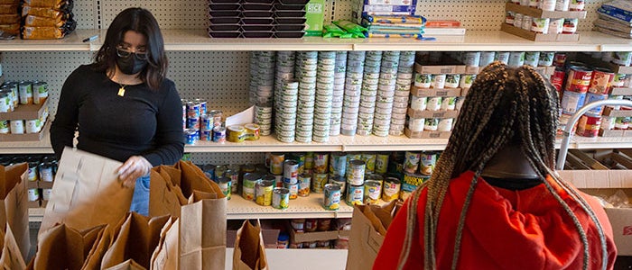 Two people are packing paper bags with various food items in a well-stocked food pantry. Shelves in the background are filled with canned goods, boxed foods, and other non-perishable items. One person is wearing a black long-sleeve shirt and a face mask, while the other has braided hair and is wearing a red hoodie.