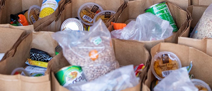 Numerous paper bags filled with various food items, including canned goods, dried beans, and plastic containers of mixed fruit, are neatly arranged in rows. The bags appear ready for distribution, with each one containing a variety of non-perishable foods. The setting suggests a food pantry or a similar community service.