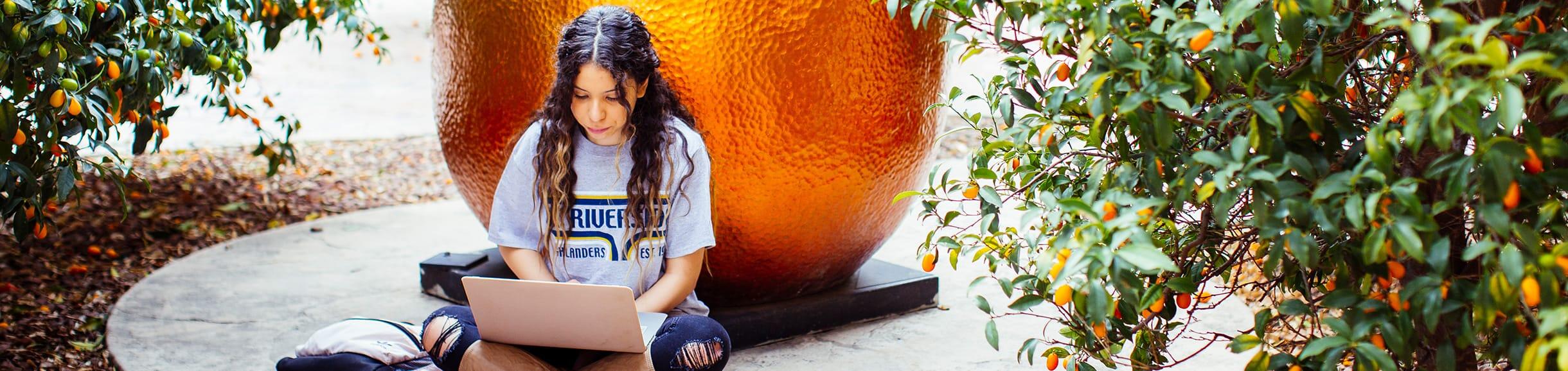 UC Riverside student sitting in front of the orange statue on her laptop